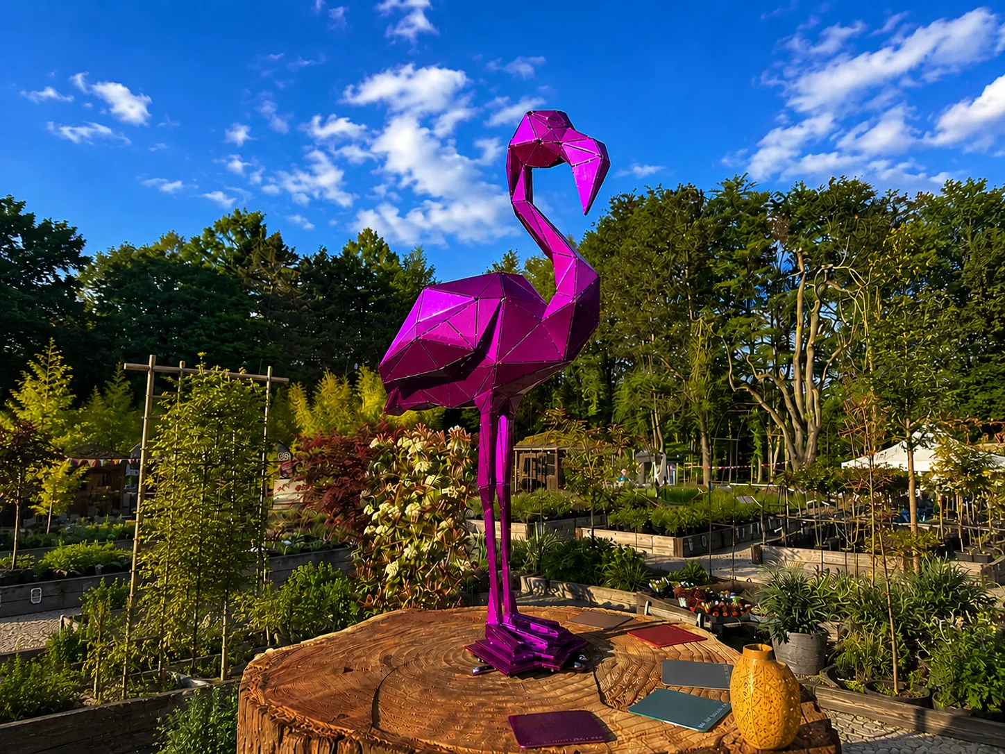 Flamingo aus Stahl in Pink auf einer Baumscheibe, bei Tag vor blauem Himmel und einer Gärtnerei